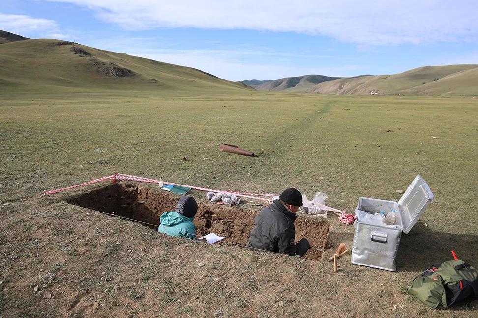 Zwei Archäologen stehen in einer Grube, in einer grünen Hügeligen Landschaft